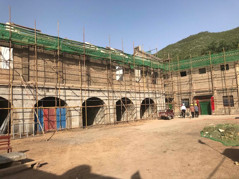 Renovation site showing arched openings wrapped in bamboo scaffolding and green mesh netting with hillside beyond