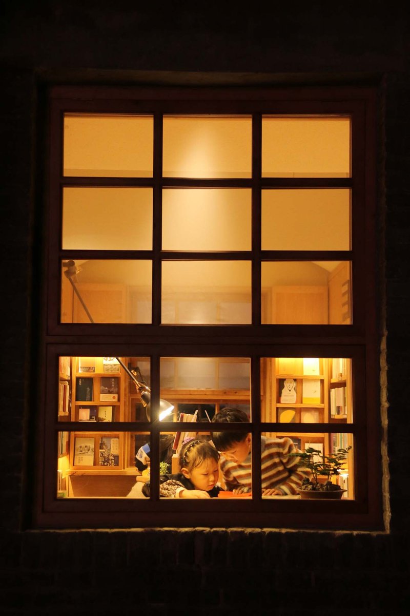 Exterior night view of a gridded window revealing two figures reading inside a warmly lit interior