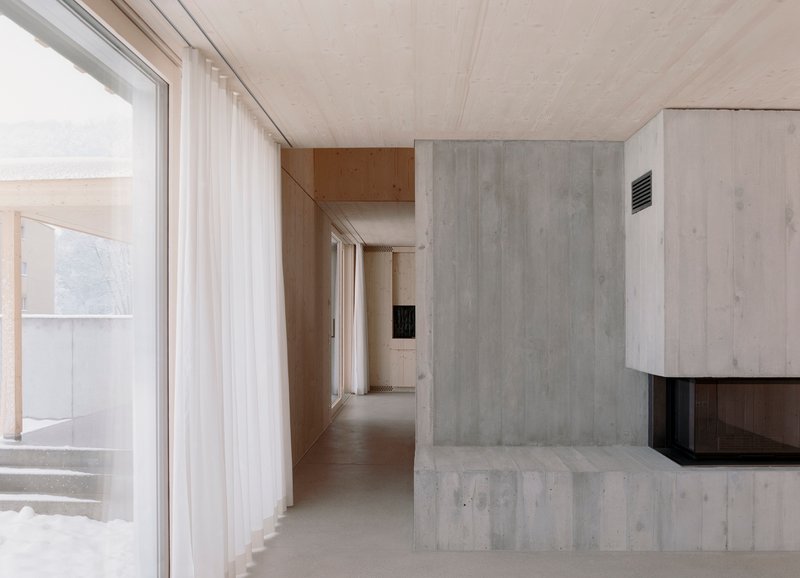 Interior corridor with exposed concrete fireplace mass, plywood ceiling, and floor-to-ceiling sheer curtains along glazing
