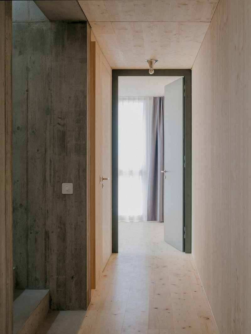 Narrow hallway with light plywood and dark timber walls leading to a room with sheer curtains