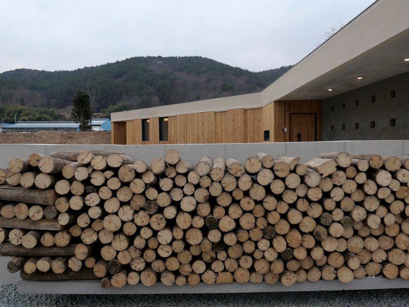 Stacked firewood logs stored in concrete planter alongside timber-clad volumes and hillside backdrop