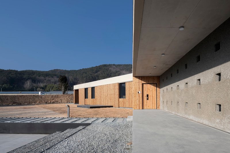 Covered walkway with concrete soffit beside gravel court and timber-clad wing under clear sky