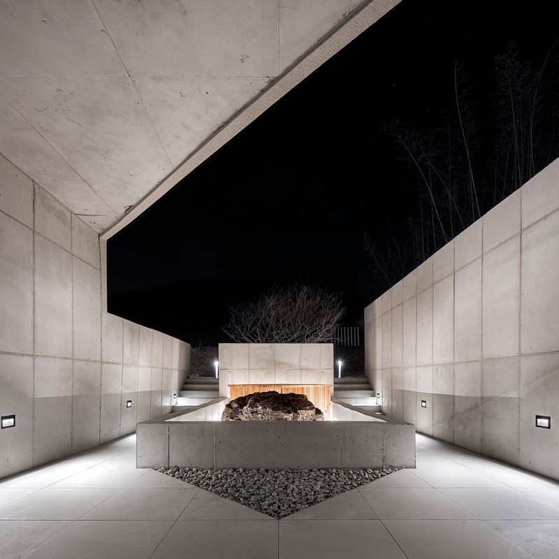 Covered entrance passage with concrete walls, overhead beams and a central planted tree at night