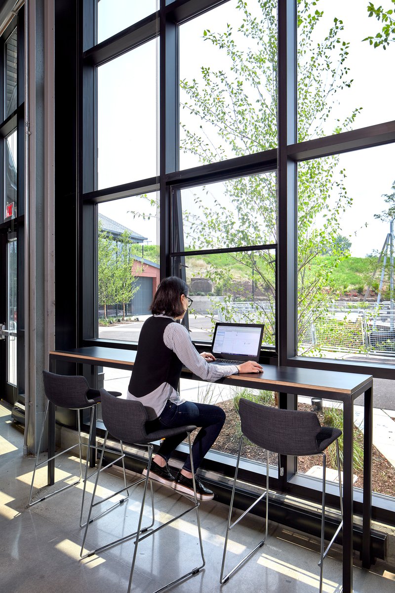 Person working at a standing-height table beside floor-to-ceiling windows overlooking exterior trees and parking lot