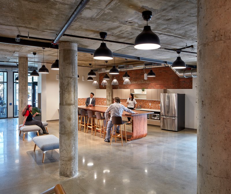 Open communal kitchen with exposed concrete columns and pendant lights as people gather at the central island