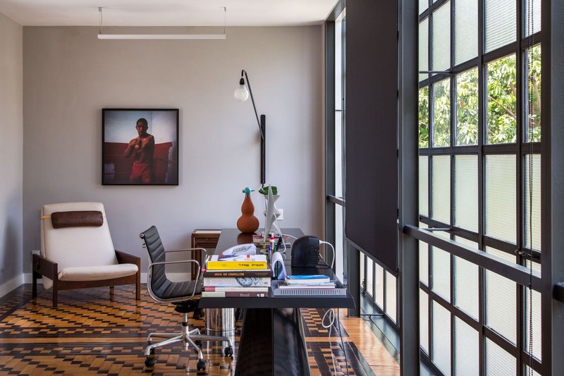 Study corner with desk and lounge chair beside tall steel-framed windows overlooking green foliage