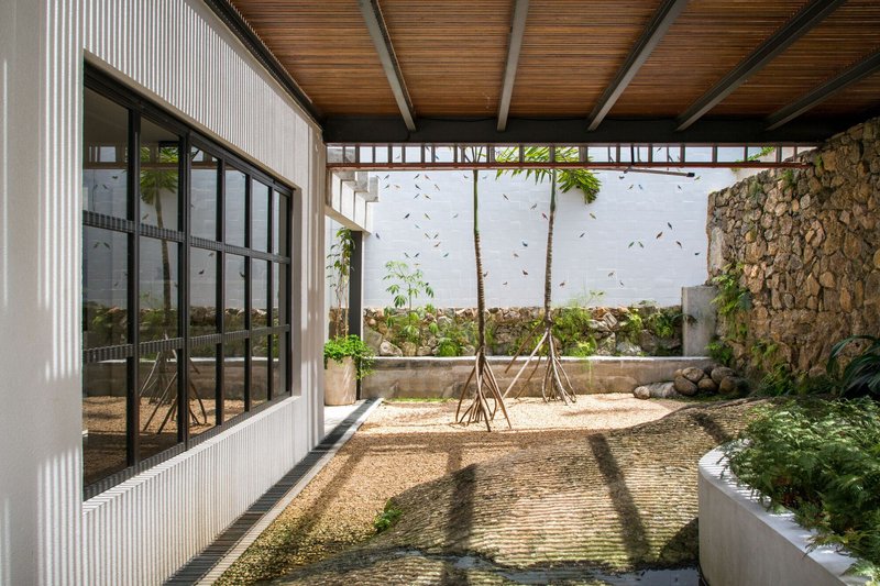 Covered courtyard with gravel floor, young trees, stone wall and timber ceiling casting afternoon shadows