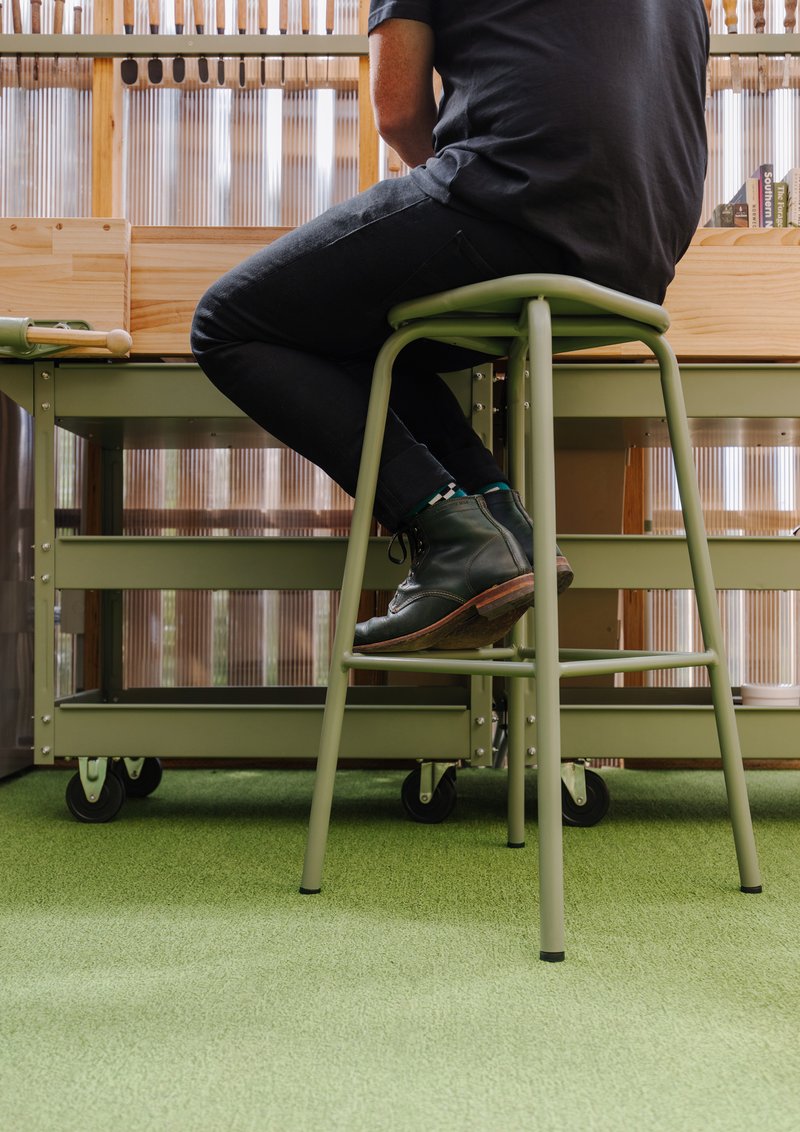 Person seated on green tubular steel stool beside rolling storage cart on artificial turf flooring