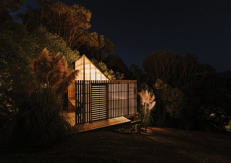 Small gabled structure with timber slat screens illuminated from within at dusk among eucalyptus trees