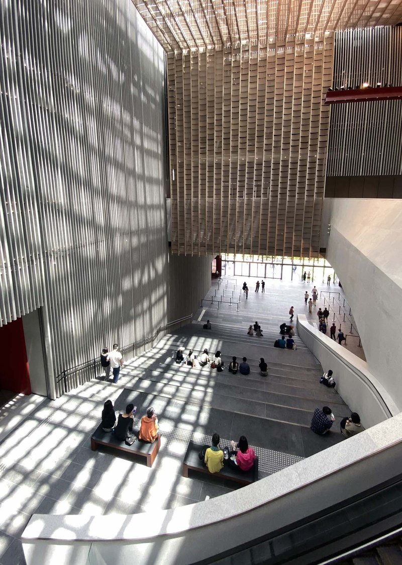 Multi-story atrium with vertical fin screens casting shadow patterns on visitors seated on steps