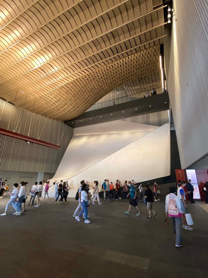 Interior lobby with vaulted timber ceiling and white sculptural wall with visitors gathered below