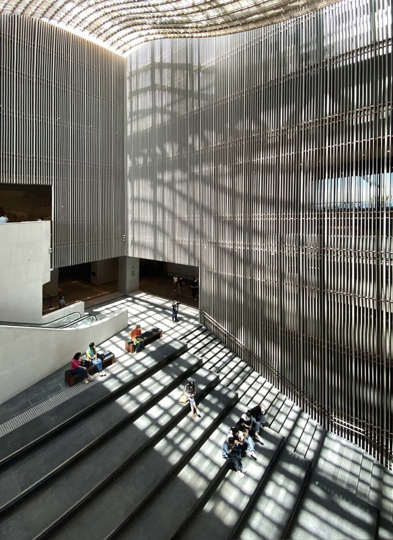 Double-height atrium with vertical metal louvers casting grid shadows on the stone floor with visitors below