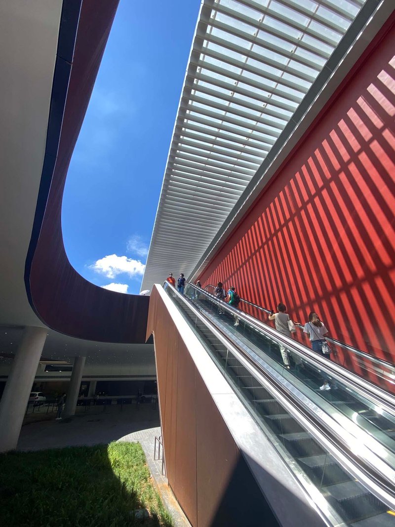 Outdoor escalator rising beneath horizontal white louvers and curved red canopy under blue sky with clouds