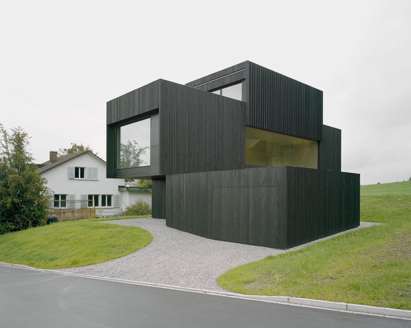 Dark timber-clad facade with stacked volumes and framed window openings on a gravel driveway