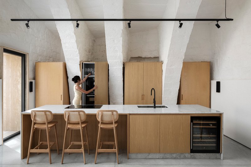 Kitchen island with light oak cabinetry and stone countertop beneath whitewashed vaulted ceiling with exposed arches