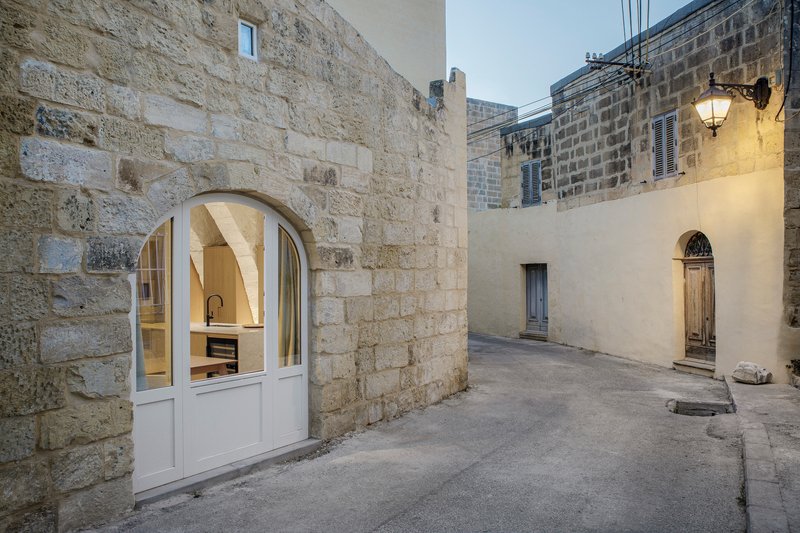 Arched doorway with white glazed frames opening onto a narrow lane between limestone block walls at dusk