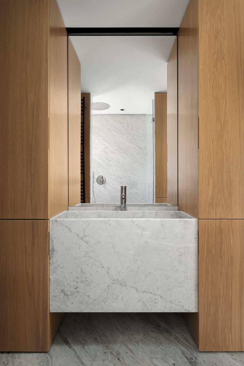 Bathroom vanity in white marble flanked by oak cabinetry with tiled shower beyond