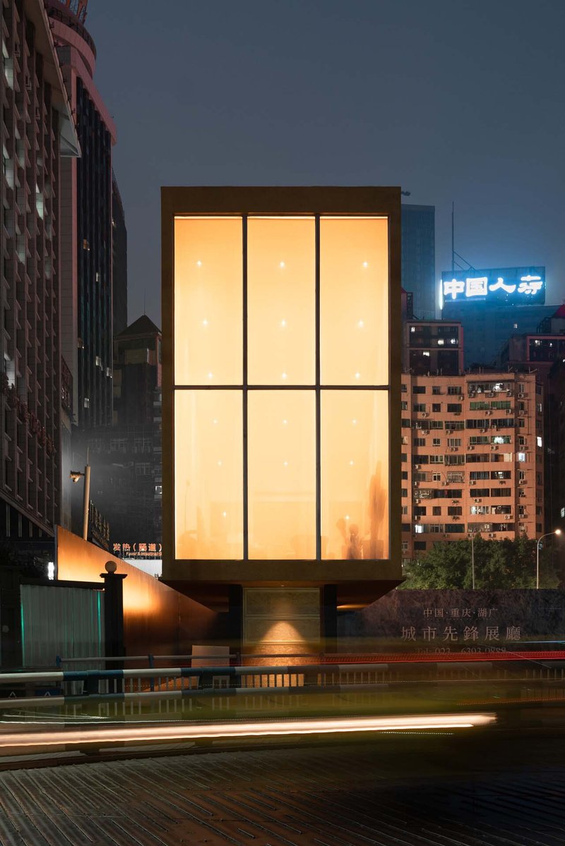 Glowing box structure with gridded translucent panels photographed at dusk with car light trails streaking past