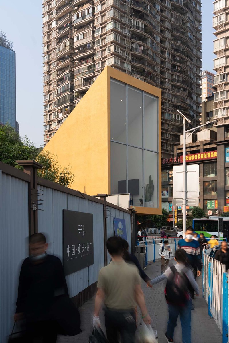 Yellow metal-clad kiosk with glazed facade elevated above street level as pedestrians pass below
