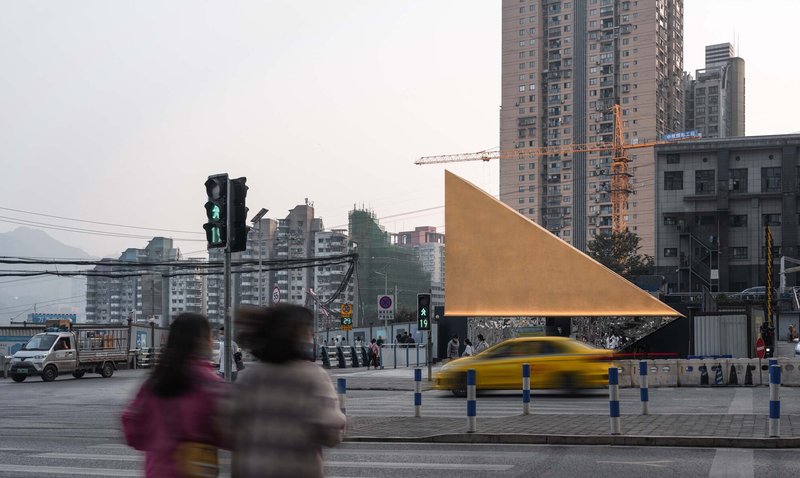 Triangular metal facade viewed from across the intersection with pedestrians and passing taxi