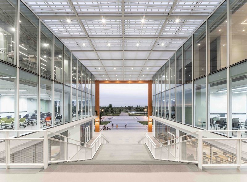 Glass-walled atrium bridge with gridded translucent ceiling panels overlooking an outdoor courtyard at twilight