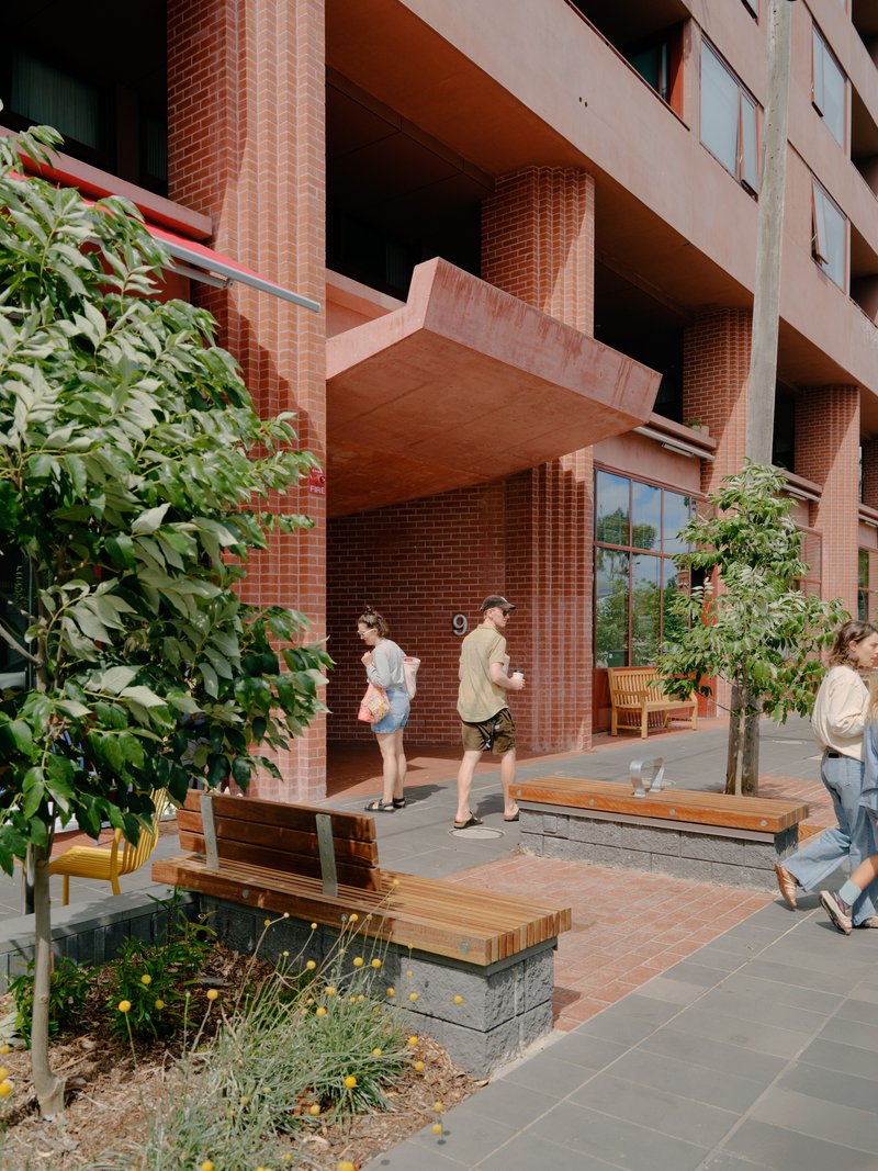 Timber bench seating beside planted beds at the base of red brick ground floor columns