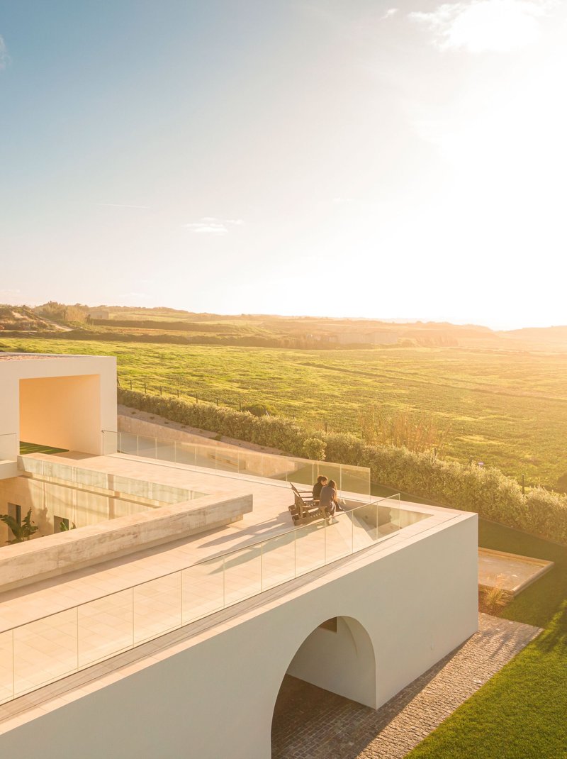 Roof terrace with glass balustrade and person seated in chair overlooking green farmland at golden hour