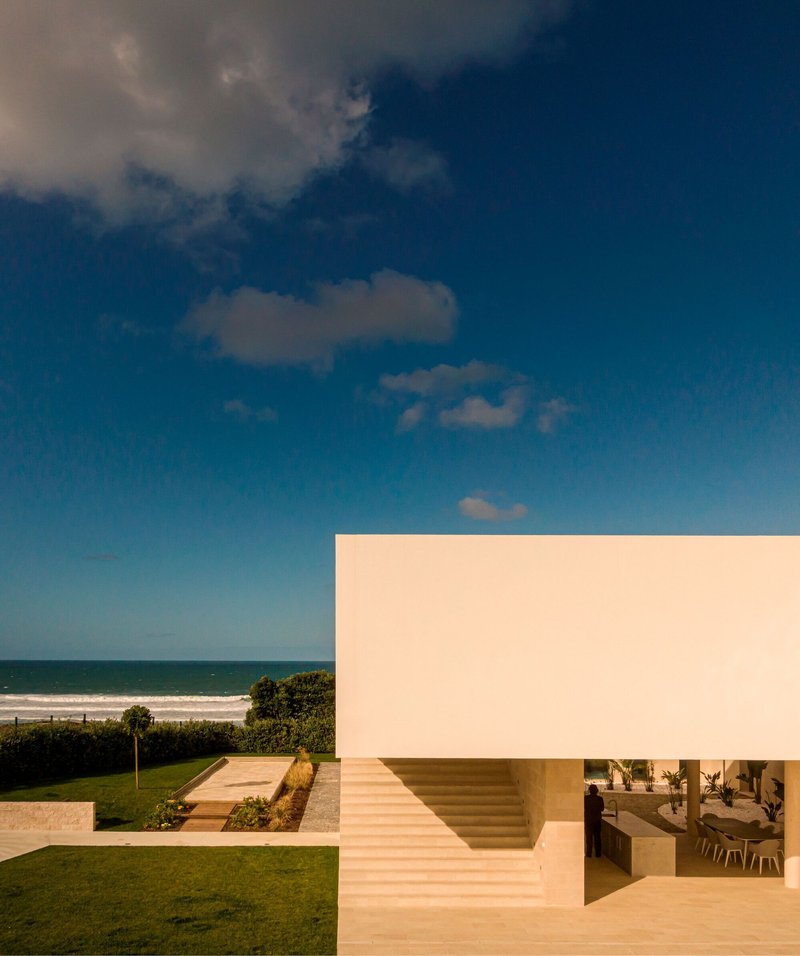 Corner view of white rendered facade with recessed staircase and ocean backdrop under blue sky