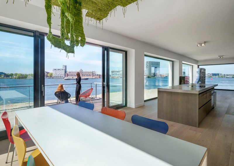 Open-plan living space with floor-to-ceiling glazing overlooking a waterfront and a hanging green textile installation