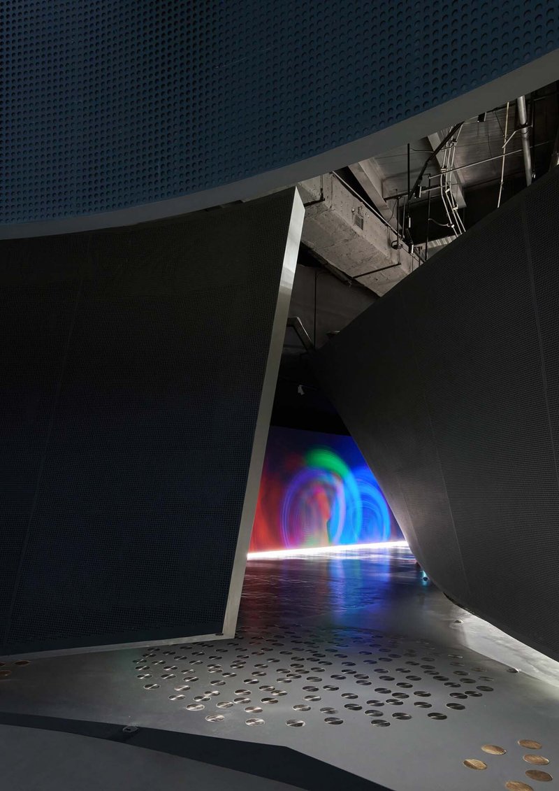 Narrow corridor with perforated walls leading to a rainbow light installation at the end