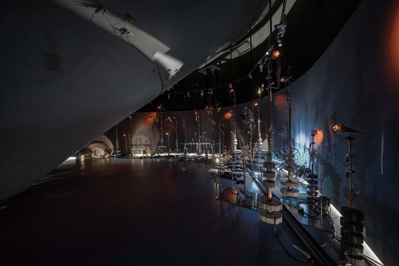 Exhibition gallery with reflective floor displaying illuminated glass sculptures along dark curved walls