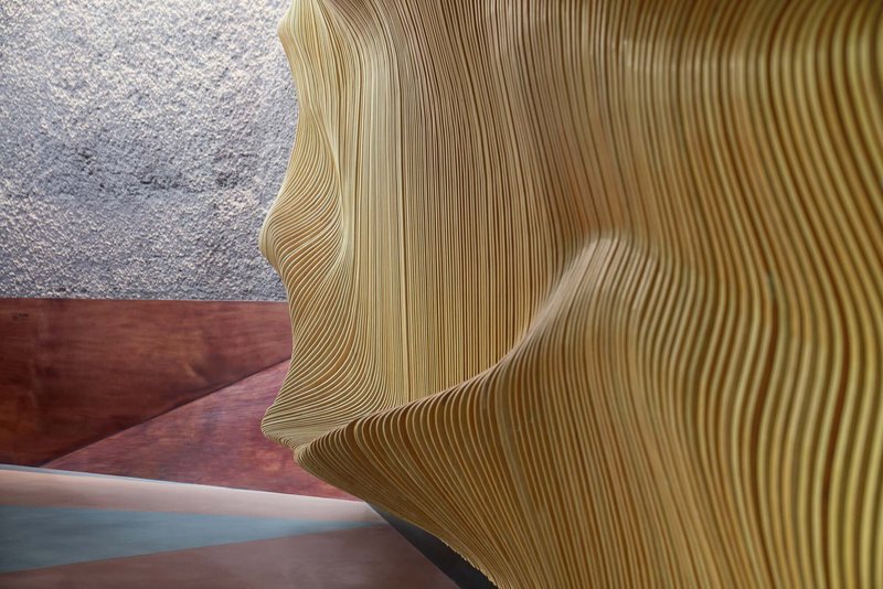 Close-up of layered wood slat wall showing undulating profile against pink floor and stone wall