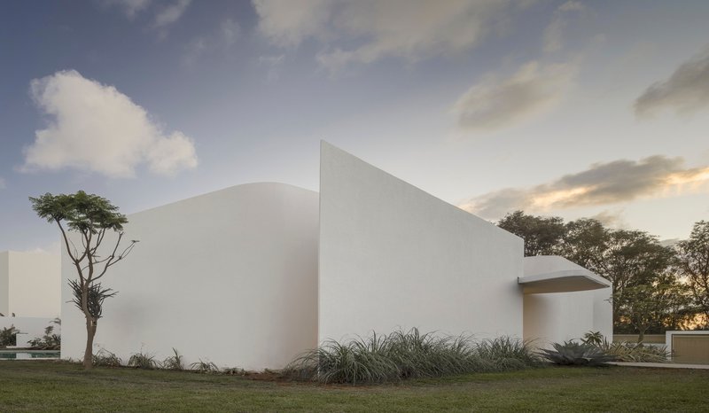 White stucco facade with curved opening flanked by grasses and a tree at dusk