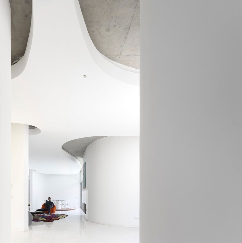 Curved white wall meeting a sculptural concrete ceiling element with a seated figure in the background