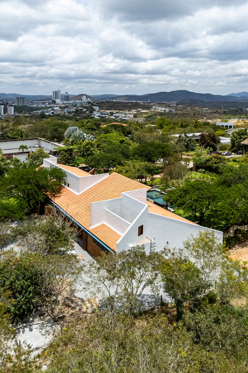 Angular orange tile roof with white parapets set among mature trees under cloudy skies