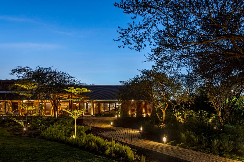 Low-slung pavilion with corrugated roof and timber screen walls along brick paved path through landscaped garden at dusk