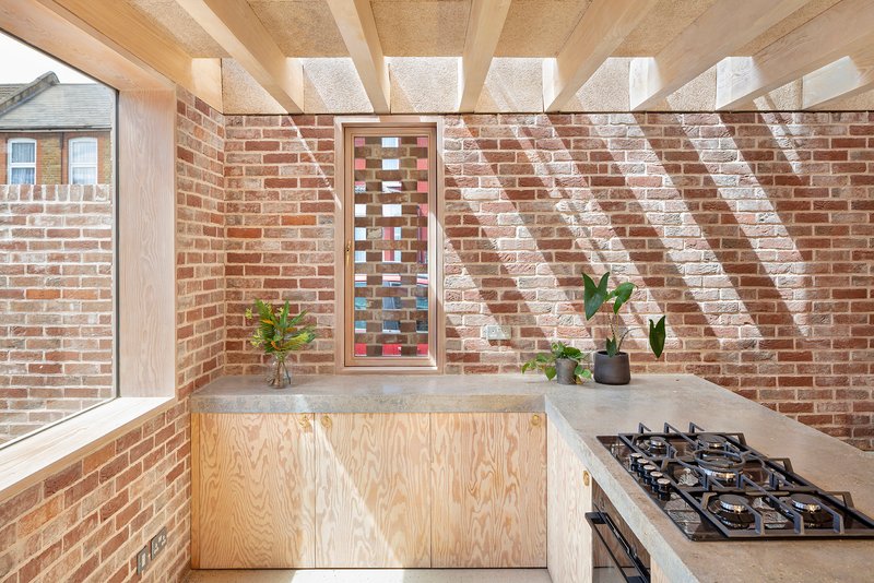 Kitchen corner with plywood cabinetry and slotted vertical window casting diagonal shadows across brick wall