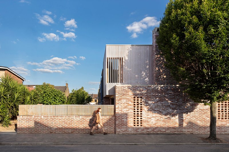 Street view of perforated brick wall with timber fence and corrugated metal upper volume as a person walks past