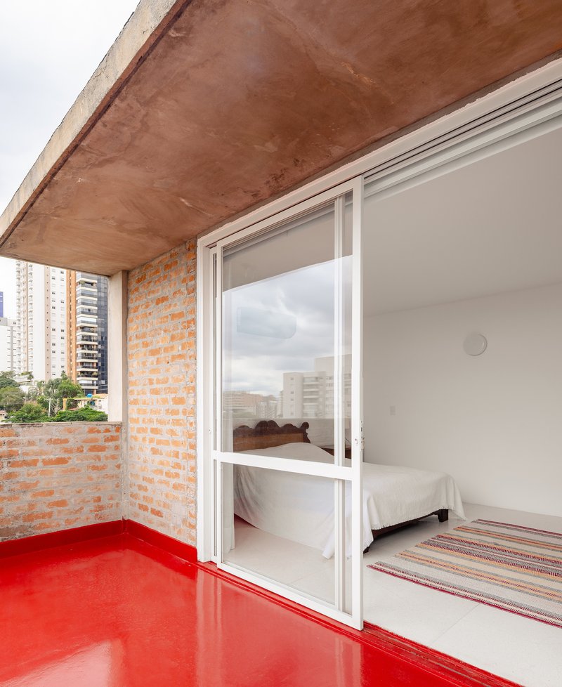 Small balcony with red resin floor and exposed brick wall opening to bedroom through sliding glass door