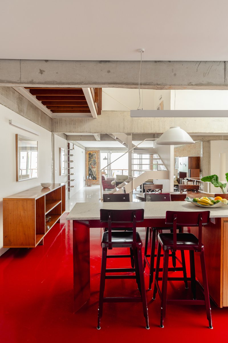 Open plan interior with exposed concrete beams, timber furniture, and bright red floor under pendant lights