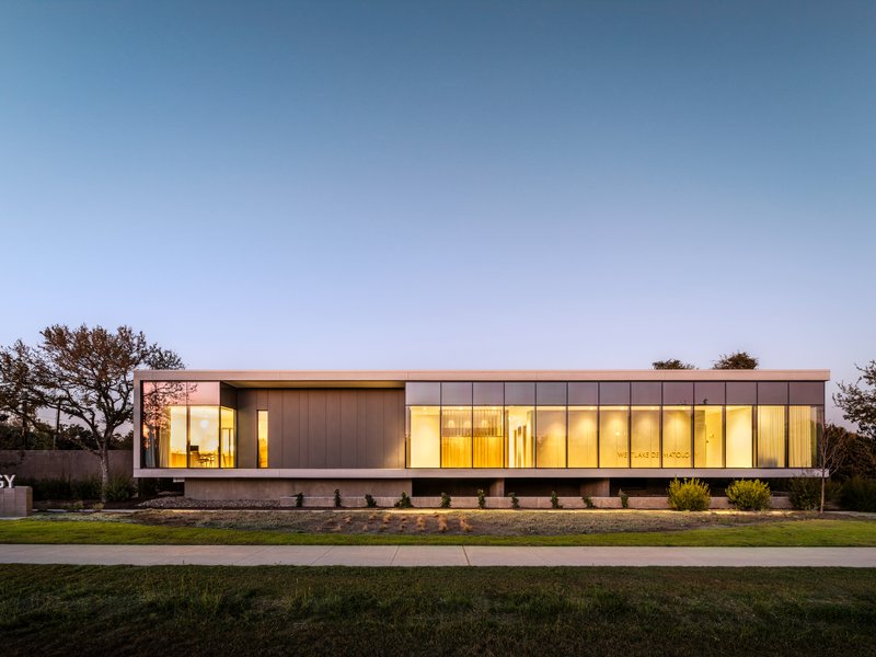 Long view of the glass and metal facade glowing at twilight behind lawn and trees