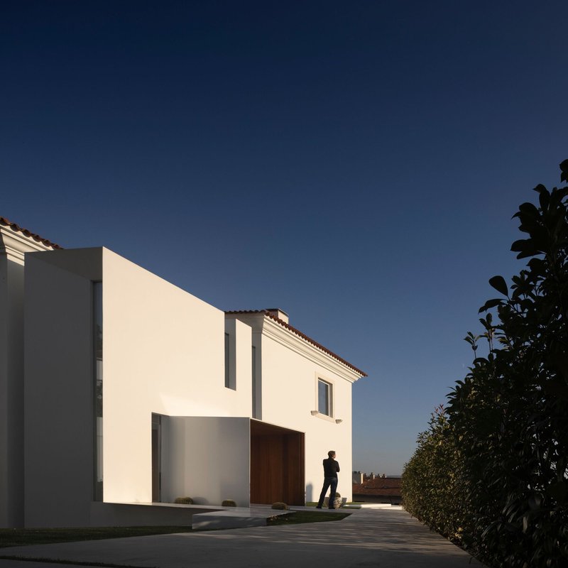 White stucco facade with recessed garage and pivoting timber door as a figure walks past at dusk
