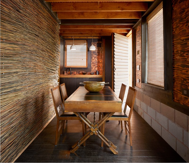 Dining area with exposed timber beams and layered wall treatments of woven screens and plywood panels