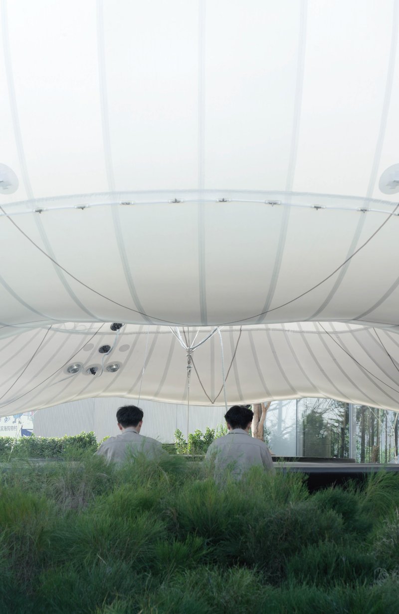 View from inside white tensioned fabric envelope looking down at two figures seated in planted grasses