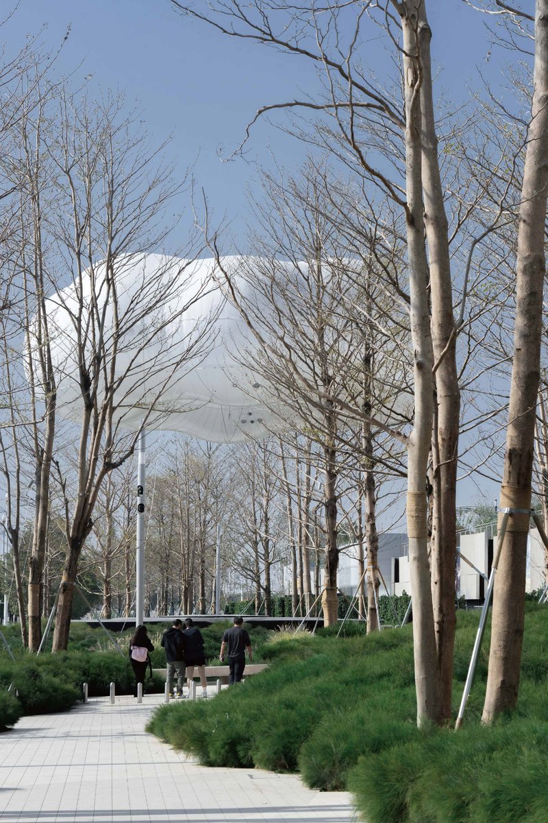 Paved pathway flanked by bare winter trees and landscaped berms leading toward a white canopy structure