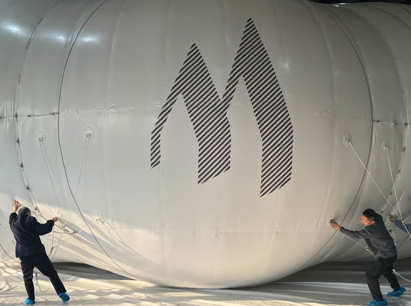 Two workers guiding ropes on a large white inflatable sphere with a striped graphic inside