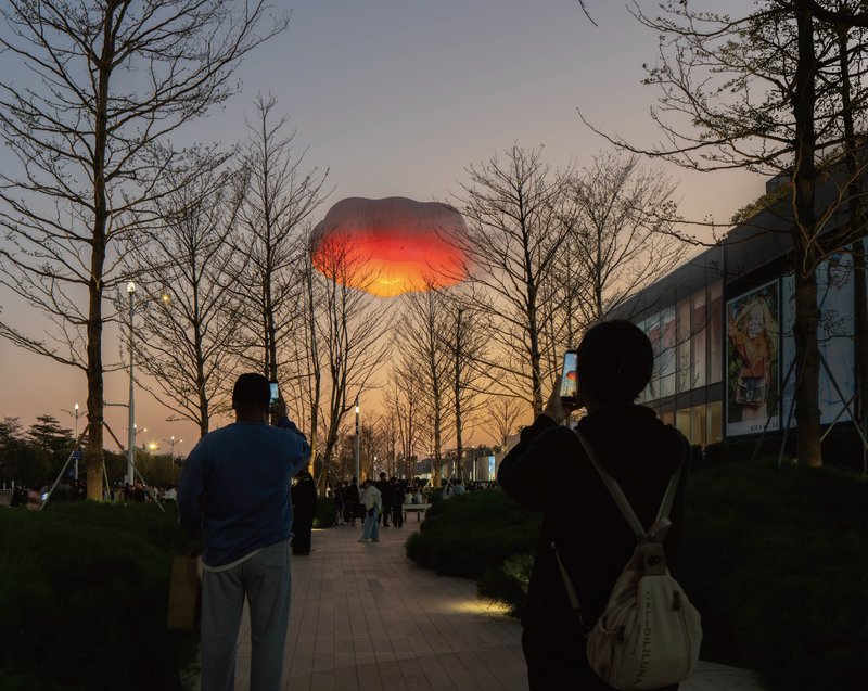 Illuminated red and orange balloon glowing at dusk above bare trees with silhouetted figures photographing below