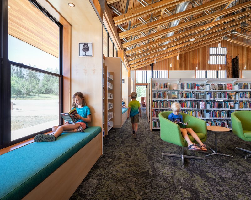 Library interior showing built-in window seat along glazed wall with children reading throughout the carpeted space