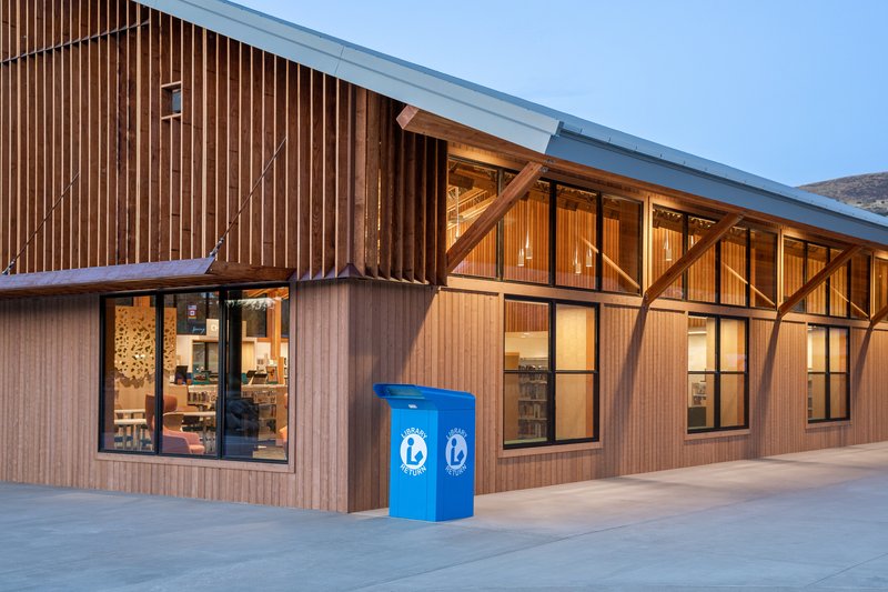 Corner view showing vertical wood cladding, exposed timber structure through glazing, and a blue recycling bin