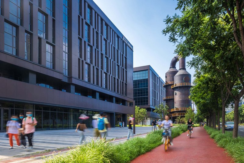 Street view of gridded facade volumes with cyclists and pedestrians on a red path beside planted beds